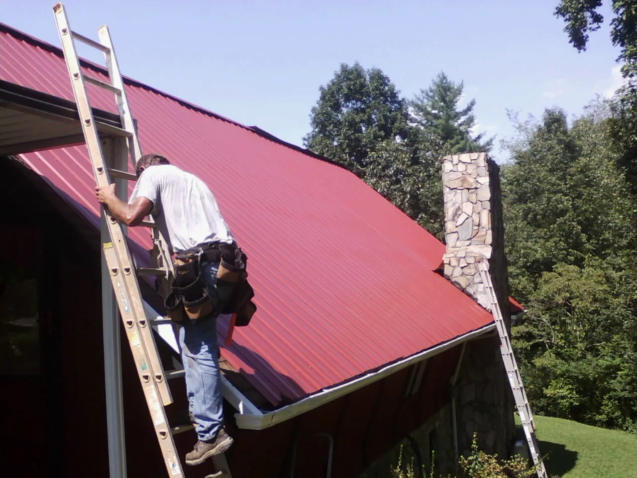 Roofer on a ladder beside a red metal roof and stone chimney — Soffit & Fascia Repair by Roofing Brothers in Stoneboro